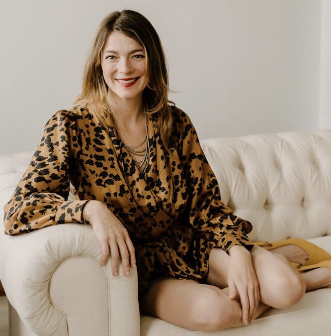 Woman with blonde hair sitting on a white sofa, wearing a leopard print dress and smiling.